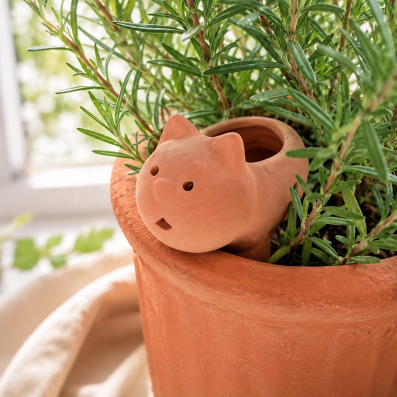 May include: A terracotta plant pot accessory shaped like a cat head with two pointed ears, two small circular eyes, and a small open mouth. The cat-shaped figure is nestled into the soil of a larger terracotta pot containing a green rosemary plant.