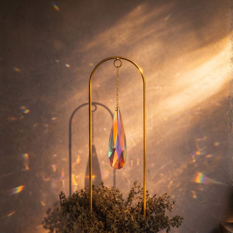 May include: A gold-colored metal stand with a teardrop-shaped crystal pendant. The crystal refracts light, displaying a spectrum of colors. The stand has an arched top and a chain suspending the crystal. Dried plants are at the base.