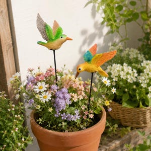 May include: Two hummingbird garden stakes featuring textured wings and bodies in vibrant shades of green, orange, yellow, and blue. Each bird is mounted on a thin black metal rod, positioned within a terracotta pot filled with colorful flowers.