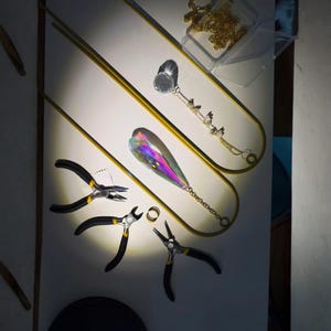 May include: Jewelry-making tools and components are arranged on a white surface. The tools include black and yellow pliers, a gold ring, and a clear plastic container with a gold chain. A large, iridescent crystal pendant and a gold chain with small charms are also present.