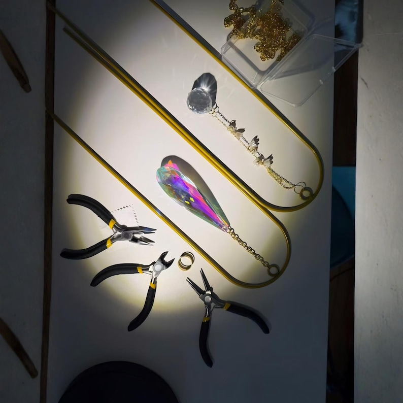 May include: Jewelry-making tools and components are arranged on a white surface. The tools include black and yellow pliers, a gold ring, and a clear plastic container with a gold chain. A large, iridescent crystal pendant and a gold chain with small charms are also present.