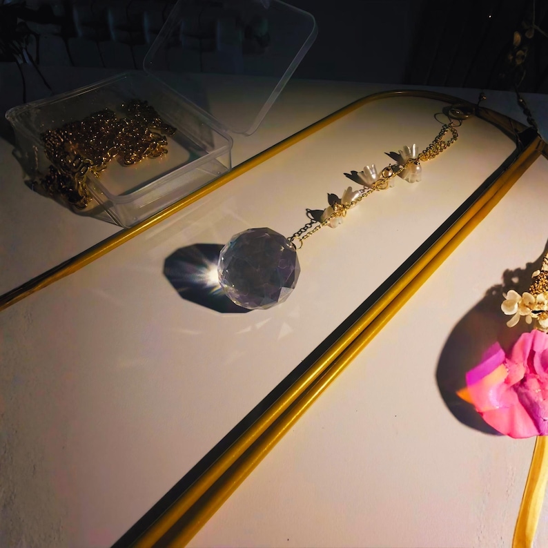 May include: A clear crystal ball pendant hangs from a gold chain, adorned with small white beads and gold accents. The pendant is suspended within a gold-colored frame, with a clear plastic container holding gold chains in the background.