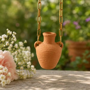 May include: A small, terracotta-colored vase with two handles, suspended by a gold-colored chain. The vase is centered in the frame, with a blurred green background of foliage and flowers. The chain is made of rectangular links.
