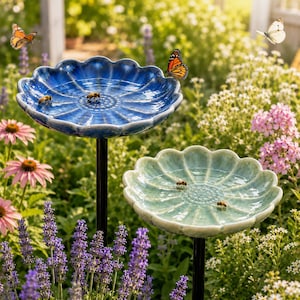 May include: Two scalloped-edge bird baths on black metal stakes, one in deep blue and one in light green, each filled with water and visited by honeybees, with a monarch butterfly perched on the blue rim and a white butterfly in the air.