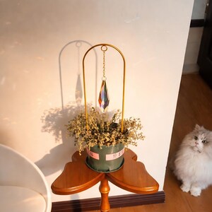 May include: A decorative table features a small green pot with dried flowers, topped with a gold-colored metal arch holding a prism-shaped crystal. The table is made of wood and has a unique, three-leaf clover shape.