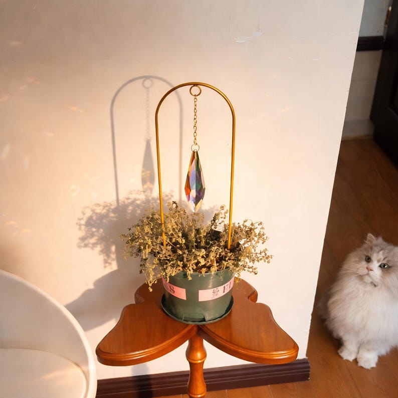 May include: A decorative table features a small green pot with dried flowers, topped with a gold-colored metal arch holding a prism-shaped crystal. The table is made of wood and has a unique, three-leaf clover shape.