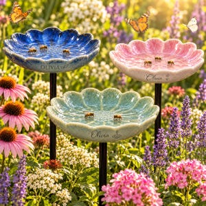 May include: Three flower-shaped bird baths in blue, pink, and light green, each mounted on a black metal stake. Each basin features a scalloped edge, a textured interior, and the name Olivia printed on the front side of the bowl.