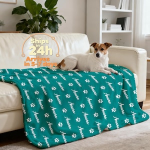 May include: A teal blanket with white paw prints and the name "Amara" repeated across the surface. A dog is lying on the blanket, which is draped over a white sofa. The blanket is a soft, cozy item.