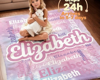 May include: A personalized plush blanket in shades of pink, purple, and blue, featuring the name "Elizabeth" in a large, stylized font. The name is repeated throughout the blanket's design. A young girl is sitting on the blanket, holding a white stuffed bunny.