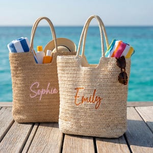 Op de afbeelding: Twee geweven strooien draagtassen met handvatten, een met "Sophia" geborduurd in roze en de andere met "Emily" in oranje. Elke tas bevat strandbenodigdheden zoals handdoeken, zonnebrandcrème en een hoed. Een zonnebril hangt aan een tas.