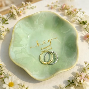 May include: A pale green ceramic ring dish with a wavy, organic edge. The center features gold lettering that reads k & g, Engaged, July.21.26. Two metal rings, one gold and one silver, rest in the center of the dish on the smooth surface.