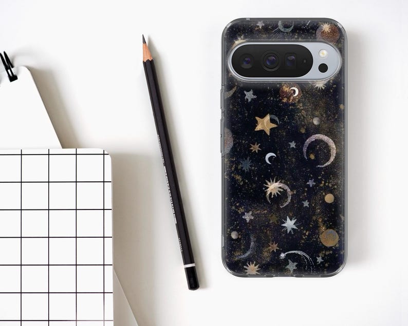 May include: A smartphone case with a celestial design featuring stars, moons, and planets in gold, silver, and white against a dark blue background. A black pencil and a white grid notebook are also in the image.