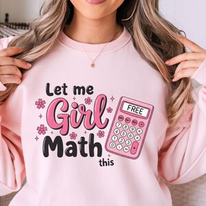以下が含まれることがあります： 「Let me Girl Math this」の文字が黒とピンクでプリントされた、薄いピンク色のスウェットシャツ。右側には「FREE」の文字が表示されたピンク色の電卓があります。ピンクの花と星もデザインに含まれています。