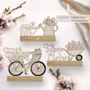 Könnte beinhalten: Dekorative Holzschilder mit Frühlingsmotiven. Eines zeigt eine Gießkanne mit Blumen, ein anderes ein Fahrrad mit einem Blumenkorb und das letzte eine Schubkarre mit Tulpen. Die Schilder stehen auf Holzsockeln.