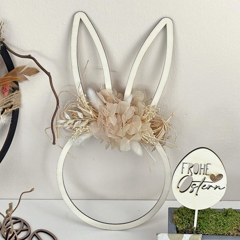 May include: A white wooden bunny rabbit shape with a wreath of dried flowers and greenery. The bunny is decorated with a wooden egg that says "Frohe Ostern" in German.