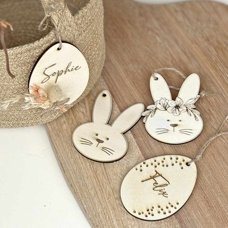 May include: Three wooden Easter egg ornaments with bunny and floral designs. One egg is personalized with the name "Sophie", another with "Felix", and the third features a bunny with a floral crown.