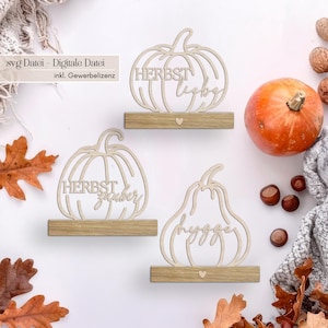 Op de afbeelding: Houten pompoenvormige decoraties met de woorden "HERBST liebe", "HERBST zauber" en "hygge" uitgesneden. De decoraties staan op kleine houten planken met hartdetails. Een oranje pompoen, noten en herfstbladeren staan ook in de afbeelding.