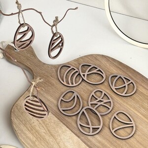 May include: Nine wooden Easter egg ornaments with a decorative cut-out design. The ornaments are brown and have a natural wood finish. They are arranged on a wooden cutting board.