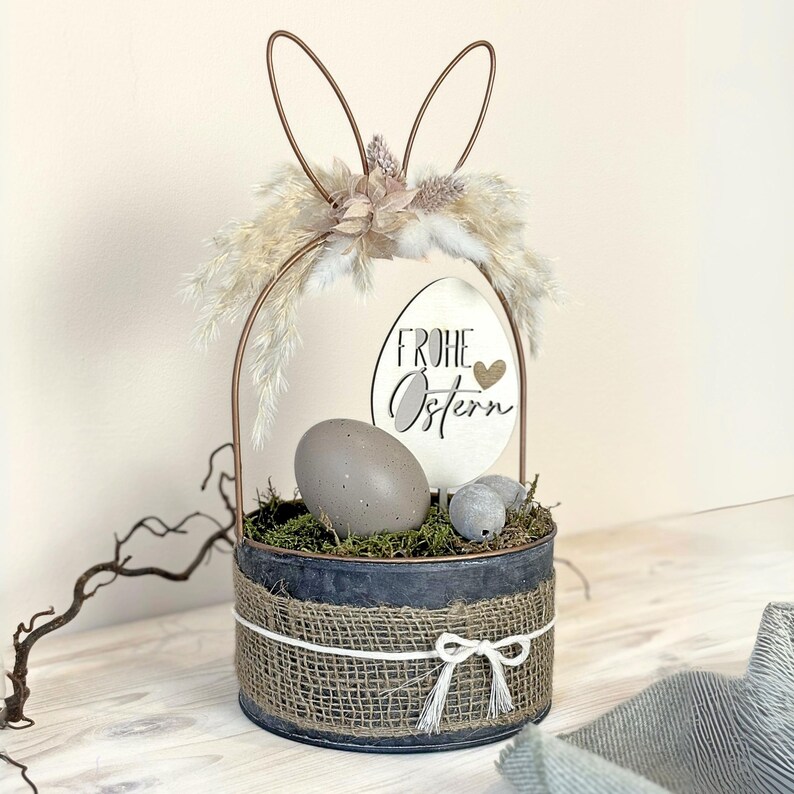 May include: A metal basket with a burlap ribbon and a wooden sign that says "Frohe Ostern" with a heart. The basket is filled with moss, faux eggs, and a fluffy white bunny tail.