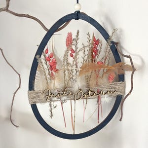 May include: A black wooden egg-shaped wreath with dried flowers and feathers. The wreath has a wooden sign that says "Frohe Ostern" which is German for "Happy Easter".