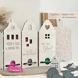 May include: Wooden tea storage boxes shaped like houses, each with a unique design and text. One reads "KEEP CALM & DRINK TEA", another "Erst Tee, dann die Welt retten", and the third "EINE TASSE Glück". The boxes have slots for tea bags and a small house-shaped decoration.