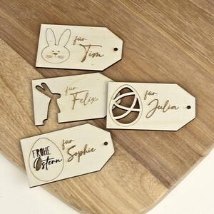 May include: Four wooden Easter gift tags with different designs. The tags are decorated with a bunny, an egg, and the words "Frohe Ostern". Each tag has a hole for string and the text "f&uuml;r" followed by a name: Tim, Felix, Julia, and Sophie.