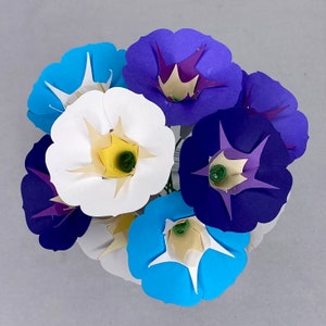 Puede incluir: Un ramo de flores de papel en tonos azules, blancos y morados. Las flores est&aacute;n dispuestas en forma circular y tienen un aspecto delicado y en capas.