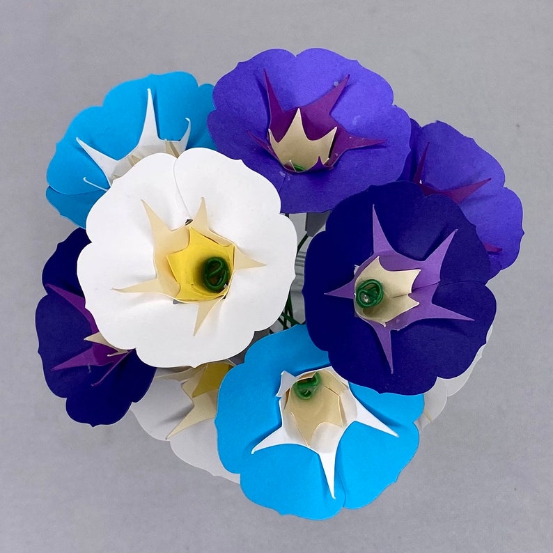 Puede incluir: Un ramo de flores de papel en tonos azules, blancos y morados. Las flores est&aacute;n dispuestas en forma circular y tienen un aspecto delicado y en capas.