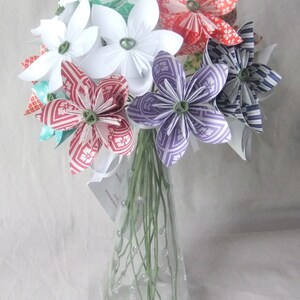 Puede incluir: Un ramo de flores de papel en un jarr&oacute;n de cristal transparente. Las flores son blancas, rojas, moradas y azules, con centros verdes. El jarr&oacute;n tiene un cuello estrecho y est&aacute; decorado con peque&ntilde;as burbujas transparentes.
