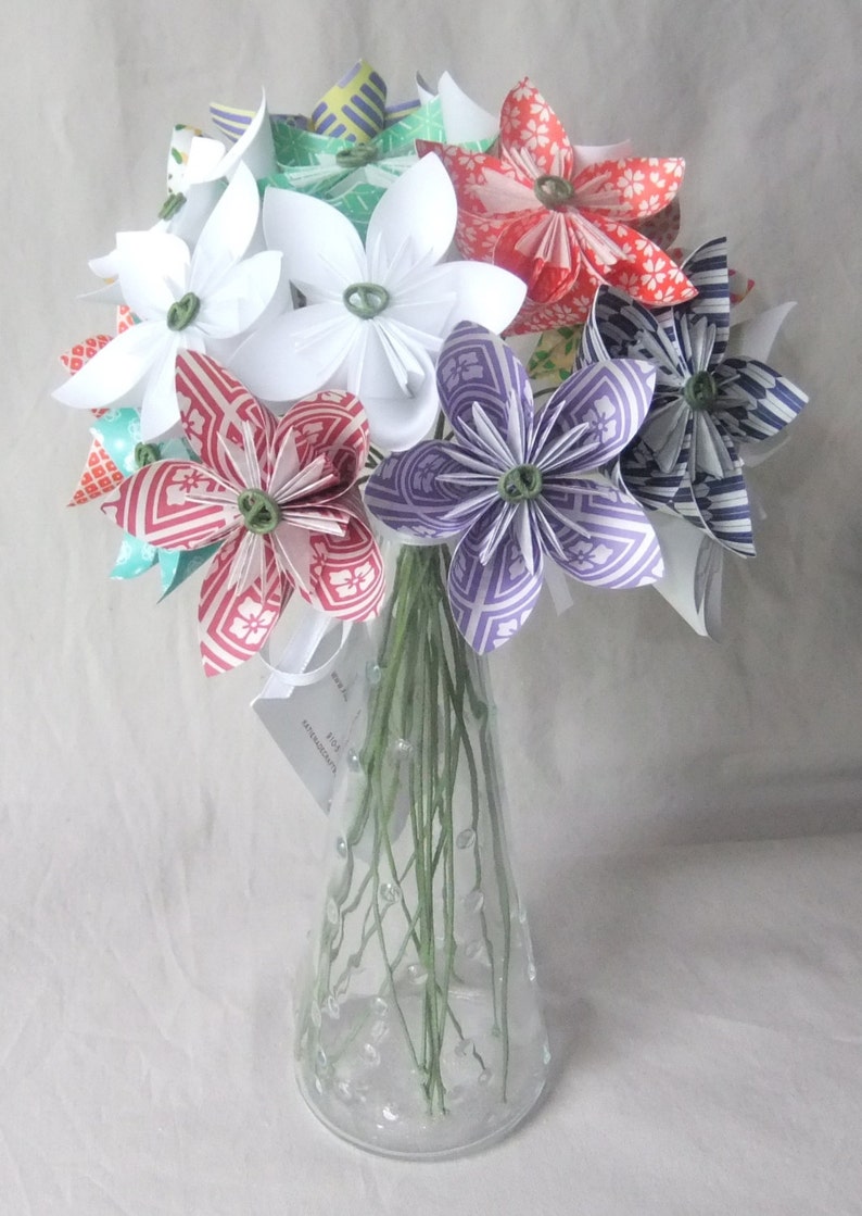 Puede incluir: Un ramo de flores de papel en un jarr&oacute;n de cristal transparente. Las flores son blancas, rojas, moradas y azules, con centros verdes. El jarr&oacute;n tiene un cuello estrecho y est&aacute; decorado con peque&ntilde;as burbujas transparentes.