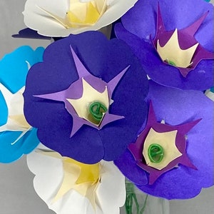 Puede incluir: Un ramo de flores de papel en tonos azules, morados y blancos. Las flores est&aacute;n dispuestas en un jarr&oacute;n y tienen un dise&ntilde;o delicado y en capas.