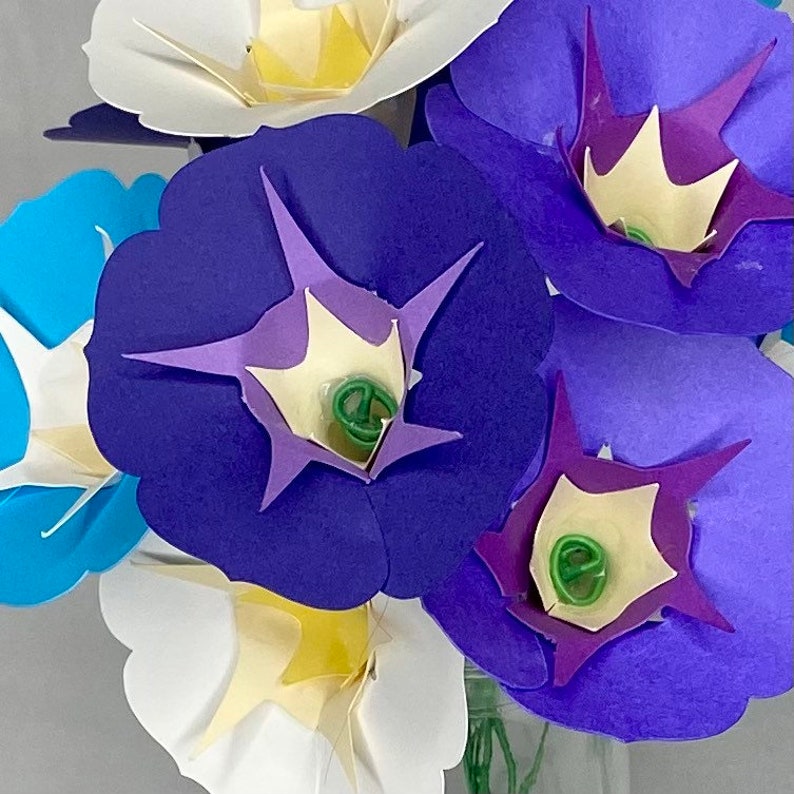 Puede incluir: Un ramo de flores de papel en tonos azules, morados y blancos. Las flores est&aacute;n dispuestas en un jarr&oacute;n y tienen un dise&ntilde;o delicado y en capas.