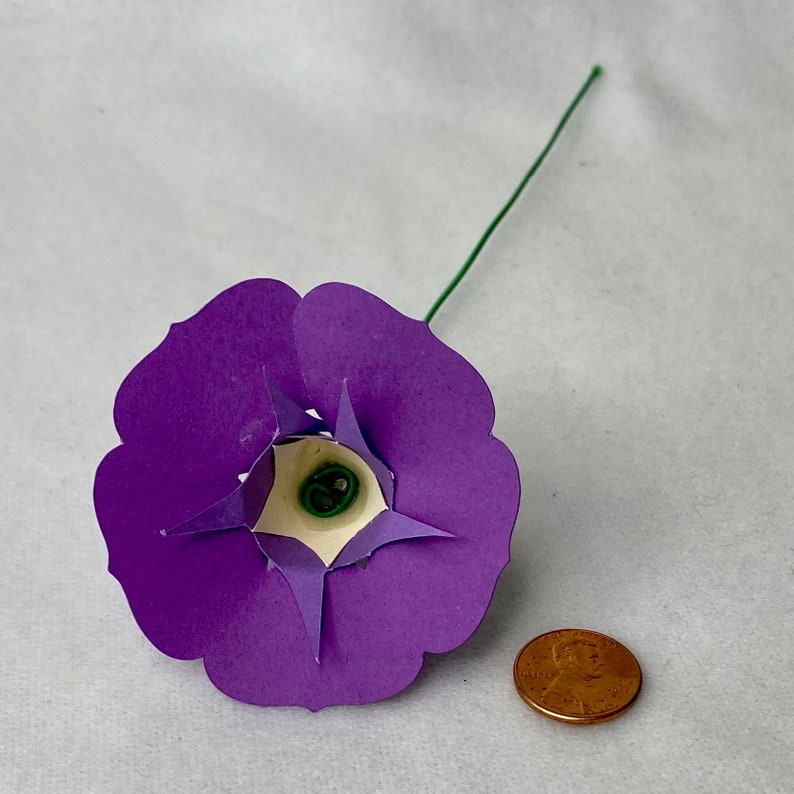 Puede incluir: Una flor de papel morada con un centro blanco y un tallo verde. La flor es aproximadamente del tama&ntilde;o de una moneda de un c&eacute;ntimo de euro.