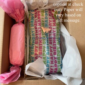 Puede incluir: Una caja de cart&oacute;n marr&oacute;n con un regalo envuelto en papel de seda rosa y blanco en su interior. El regalo est&aacute; envuelto en papel de colores con las palabras "&iexcl;Feliz cumplea&ntilde;os!" repetidas. Una peque&ntilde;a mariposa rosa est&aacute; pegada al papel de regalo.