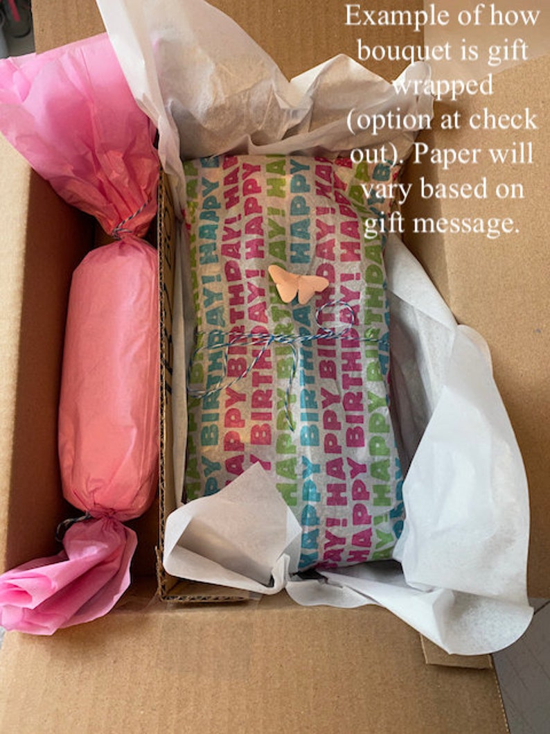 Puede incluir: Una caja de cart&oacute;n marr&oacute;n con un regalo envuelto en papel de seda rosa y blanco en su interior. El regalo est&aacute; envuelto en papel de colores con las palabras "&iexcl;Feliz cumplea&ntilde;os!" repetidas. Una peque&ntilde;a mariposa rosa est&aacute; pegada al papel de regalo.