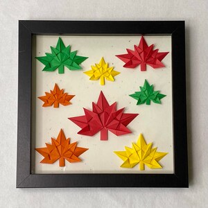 May include: A framed artwork featuring eight origami leaves in various shades of red, orange, yellow, and green. The leaves are arranged in a symmetrical pattern on a white background.