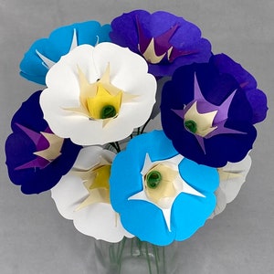 Puede incluir: Un ramo de flores de papel en tonos azules, morados y blancos. Las flores est&aacute;n dispuestas en un jarr&oacute;n y tienen un aspecto delicado, similar al origami.