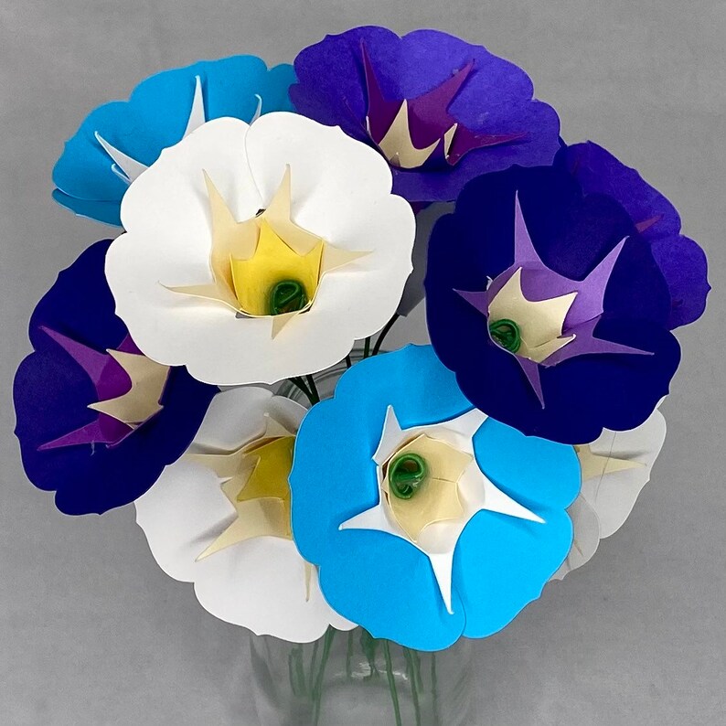 Puede incluir: Un ramo de flores de papel en tonos azules, morados y blancos. Las flores est&aacute;n dispuestas en un jarr&oacute;n y tienen un aspecto delicado, similar al origami.