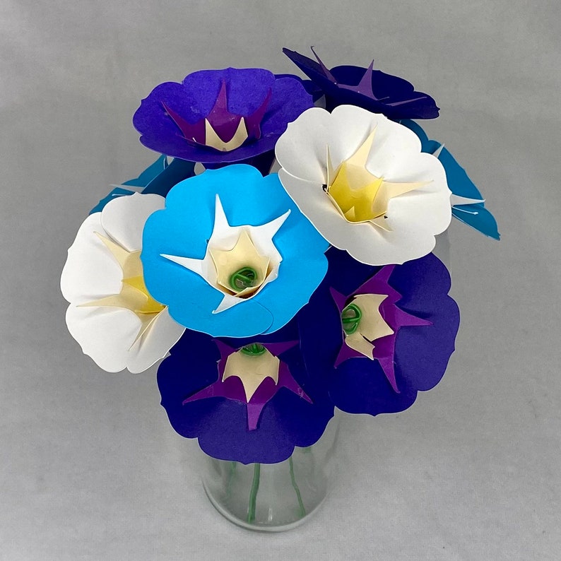 Puede incluir: Un ramo de flores de papel en un jarr&oacute;n de vidrio. Las flores son azules, moradas y blancas, y est&aacute;n dispuestas en un estilo suelto y natural.