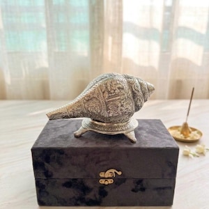 May include: A silver-toned conch shell featuring intricate embossed carvings of figures and floral patterns, resting on a matching three-legged metal stand. The shell is positioned atop a dark gray rectangular velvet box with a gold-colored latch.