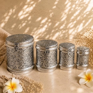 Peut inclure: Un ensemble de quatre boîtes cylindriques en métal argenté de tailles variées, chacune présentant un motif floral en relief complexe, une bordure texturée en corde en haut et en bas, et un petit fermoir à charnière sur le devant du couvercle.