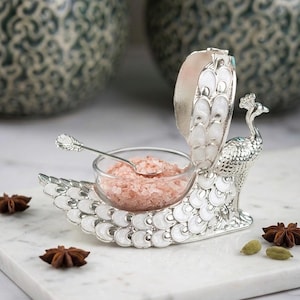 May include: A silver-toned peacock-shaped condiment holder with white enamel detailing on the tail feathers. The piece features a small, clear glass bowl filled with pink salt crystals and includes a matching silver-toned metal spoon.