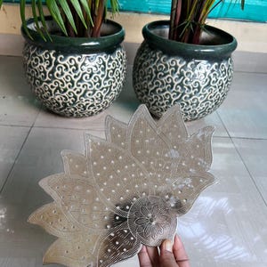 May include: A silver-coloured, lotus-shaped decorative tray with intricate cut-out designs. The tray is held in a hand. Two ceramic pots with green rims and white and green swirl patterns are in the background, each containing a green plant.