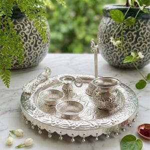 May include: A silver-toned metal puja thali set featuring a circular tray with embossed floral patterns and hanging bells. The set includes a small peacock figurine, two shallow bowls, a tall incense holder, and a rounded pot with intricate carvings.