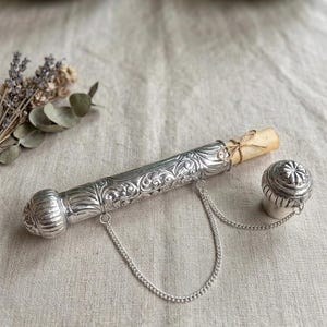 Peut inclure: Un étui à parchemin en métal argenté orné d'un motif floral en relief sur tout le corps cylindrique. L'objet comprend un capuchon amovible assorti relié par une fine chaîne argentée et contient un parchemin roulé attaché avec de la ficelle.