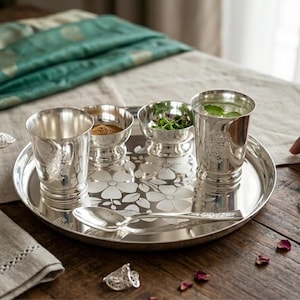 Puede incluir: Un juego de servir en tono plateado pulido que consta de una bandeja redonda con un patrón floral grabado, dos vasos a juego, dos cuencos pequeños y una cuchara. La bandeja presenta un diseño floral central, mientras que los vasos y la cuchara tienen detalles en relieve.