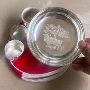 Op de afbeelding: Een zilveren puja thali set met een gedetailleerd gegraveerd ontwerp van een koe en kalf onder een boom. De set bevat een ronde plaat en drie kleine bekers, rustend op een rode etui met ritssluiting.