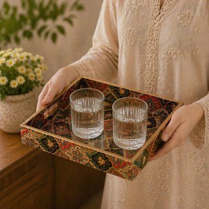 May include: A rectangular wooden tray with a detailed, patterned design in red, green, and gold. Two clear, ribbed glasses filled with water sit on the tray. The tray is held by a person wearing a beige top.
