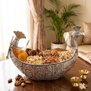 May include: A silver-toned metal serving bowl shaped like a boat with a peacock head at one end and a tail at the other. The exterior features intricate embossed floral patterns. It is divided into four sections filled with dried fruits and nuts.