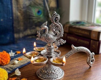 May include: A silver-toned metal oil lamp featuring a central peacock figure atop a pedestal base. The lamp has five small bowls for wicks, each holding a lit flame, and a curved handle extending from the side, all decorated with intricate patterns.